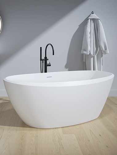 Baignoire en surface solide blanche gia de fleurco. La baignoire a une forme ovale symétrique et est blanche mate. Il y a une lueur de soleil spectaculaire provenant d'une fenêtre hors du cadre de l'image. Le mur est bleu et le sol est en lattes de bois.
