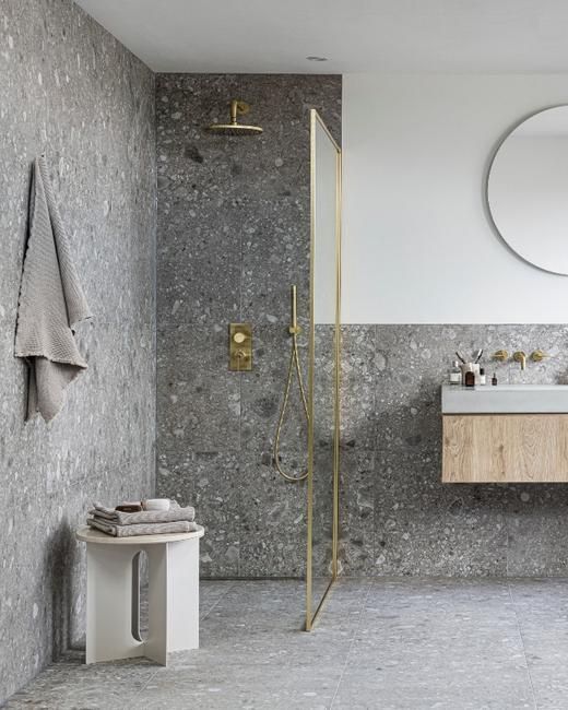Une salle de bains élégante avec une douche, un lavabo et un banc en bois. Les murs en pierre grise et les touches d'or brossé ajoutent une touche sophistiquée.